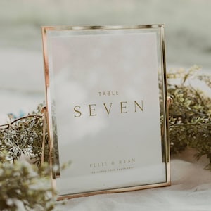 Może przedstawiać: Złota ramka z numerem stołu z numerem "SEVEN" w złotych literach. Znak zawiera również imiona "Ellie & Ryan" i datę "Saturday 18th September". Znak jest wyświetlany na białej powierzchni z zielenią na pierwszym planie.