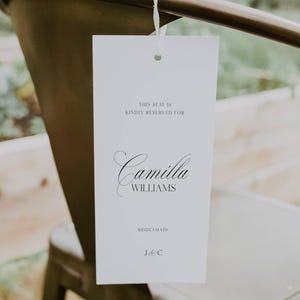 May include: A white rectangular wedding seating card with the text "THIS SEAT IS KINDLY RESERVED FOR" above the name "Camilla Williams". The word "Bridesmaid" is below the name. The card is tied to a metal chair.