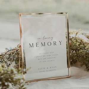 以下が含まれることがあります： 金色の金属フレームに入った看板。看板には「in loving MEMORY」と書かれており、その下にテキストが追加されています。看板は緑に囲まれ、白い表面に置かれています。テキストには名前と日付が含まれています。