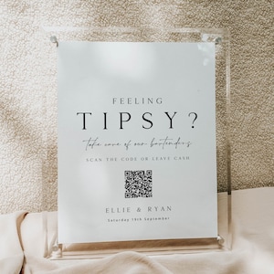 May include: A white sign with black text that reads "Feeling Tipsy? Take care of our bartenders. Scan the code or leave cash." A black and white QR code is below the text. The sign also includes the names "Ellie & Ryan" and the date "Saturday 19th September".