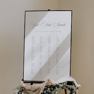 May include: A large white seating chart with black text, displayed on a wooden easel. The text reads "Your Seat Awaits" and lists names and table assignments for Juliette & Charles' wedding on August 28, 2026. The easel is decorated with greenery and fabric.