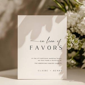 May include: A white card with the text "in lieu of FAVORS" in a stylish font. The card also includes the text "in lieu of traditional wedding favors, we have made a donation to THE AMERICAN CANCER CHARITY" and the names "CLAIRE • HENRY". White flowers are in the background.