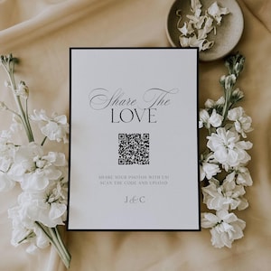 Puede incluir: Un letrero blanco con borde negro, con el texto "Share The LOVE" y un código QR. El letrero está rodeado de flores blancas y descansa sobre una superficie beige. La parte inferior del letrero dice "SHARE YOUR PHOTOS WITH US! SCAN THE CODE AND UPLOAD" y "J & C".