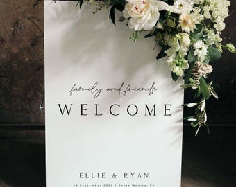 May include: White welcome sign with black text that reads "family and friends welcome Ellie & Ryan 19 September 2023 | Santa Monica, CA". The sign is decorated with white flowers.