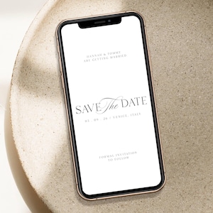 Op de afbeelding: Een smartphone toont een digitale huwelijksuitnodiging met de tekst "SAVE The DATE" in elegant schrift. De uitnodiging bevat de namen "HANNAH & TOMMY" en de datum "03.09.26 VENICE, ITALY". De telefoon heeft een roségouden frame en ligt op een neutrale ondergrond.