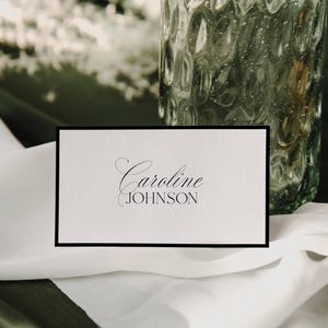 Może przedstawiać: Biała wizytówka z czarną obwódką, z imieniem i nazwiskiem "Caroline Johnson" w eleganckiej czcionce. Karta spoczywa na białej tkaninie, z teksturowanym szklanym wazonem w tle.