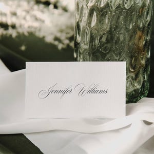 May include: A white place card with the name "Jennifer Williams" in elegant script. The card rests on a white cloth, with a textured glass vase in the background. The overall aesthetic is simple and refined.