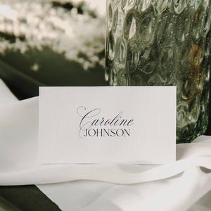 Puede incluir: Una tarjeta de lugar blanca con el nombre "Caroline Johnson" impreso en una elegante escritura negra. La tarjeta descansa sobre una tela blanca, con un jarrón de vidrio texturizado en el fondo.