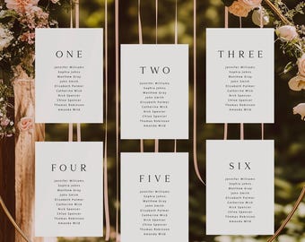 May include: A gold metal hoop adorned with flowers and greenery, with six white cards hanging inside. Each card is labeled with a number and lists names, likely for a wedding seating arrangement. Ribbons and floral arrangements add to the decorative design.