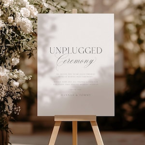 Puede incluir: Un letrero blanco sobre un caballete de madera con las palabras "UNPLUGGED Ceremony". El letrero también incluye el texto "Te invitamos a estar plenamente presente con nosotros durante nuestra ceremonia. Gracias. Hannah & Tommy."