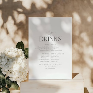 May include: A white rectangular sign with the words "The Drinks" in elegant script. Below, a list of wine, beer, spirits, soft drinks, and signature cocktails is displayed in black text. A white floral arrangement is in the lower left.
