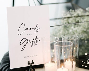 May include: A white sign with black calligraphy that reads "Cards and Gifts" with the words "Thank You!" underneath. The sign is on a black stand and is sitting on a white marble table with a glass box and candles.