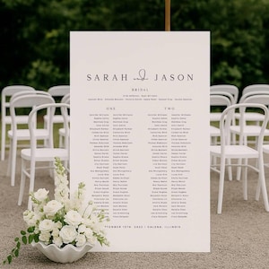 Peut inclure: Un plan de table de mariage blanc avec les noms "Sarah & Jason" en haut. Les noms des invités sont listés sous les colonnes "ONE" et "TWO". Un arrangement floral de roses blanches et de verdure est devant le panneau. Des chaises blanches sont en arrière-plan.