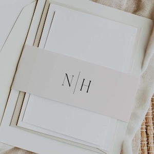 May include: A white wedding invitation with a blush pink band that has the initials "N|H" in black.