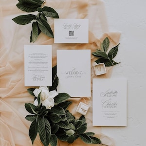 May include: Elegant wedding invitation suite with white cards, featuring the text "The Wedding" and names. Includes a "Kindly Reply" card, details card, and a card with names. Embellished with greenery and rings.