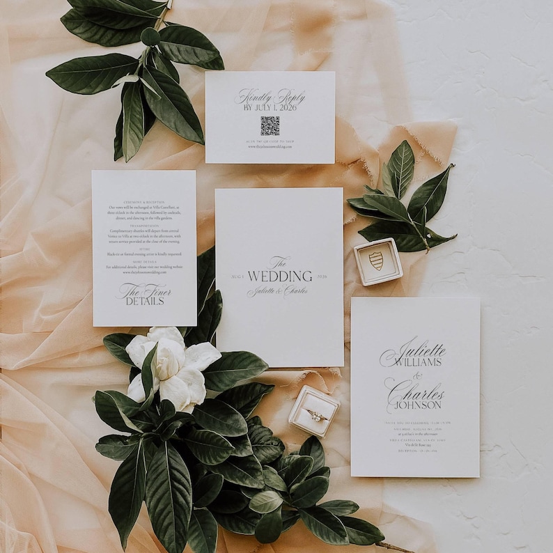 May include: Elegant wedding invitation suite with white cards, featuring the text "The Wedding" and names. Includes a "Kindly Reply" card, details card, and a card with names. Embellished with greenery and rings.