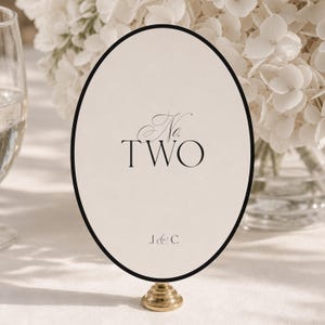 Puede incluir: Un letrero ovalado para número de mesa con fondo color crema y un borde negro fino. El texto dice No. TWO en una combinación de fuentes script y serif, con las iniciales J&C impresas debajo. El letrero está montado sobre una base dorada.