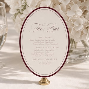 May include: An oval-shaped bar menu sign with a cream background and a dark burgundy border, mounted on a small gold-toned pedestal base. The text reads The Bar, Wine, Beer, Spirits, Soft Drinks, and Signature Cocktails with listed drink options.