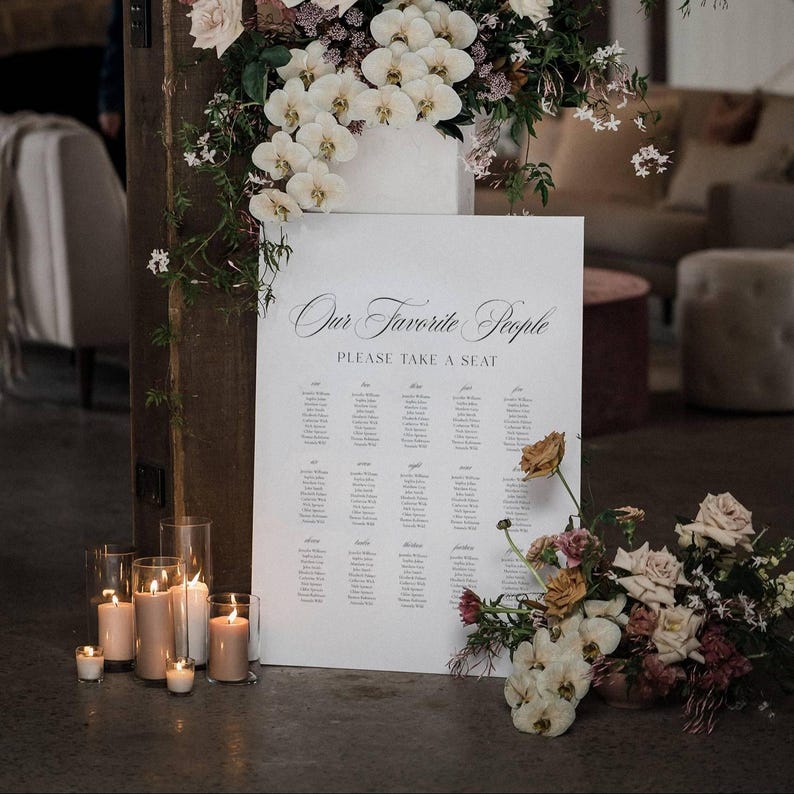 May include: A white sign with the text "Our Favorite People" and "Please Take a Seat" in elegant script. The sign lists names, likely for a wedding or event. The scene includes candles, flowers, and a floral arrangement.