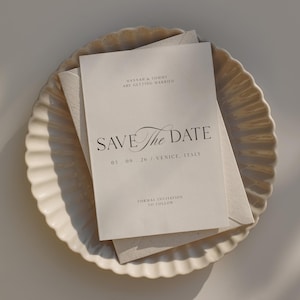 Op de afbeelding: Een crèmekleurige trouwkaart met de tekst "SAVE The DATE" en de datum "03.09.26 VENICE, ITALY". De uitnodiging ligt op een geschulpte schaal met een envelop.