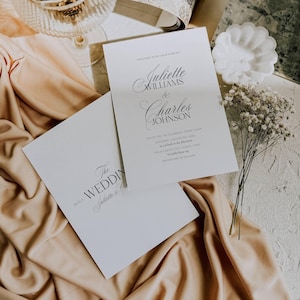 May include: Two white wedding invitation cards with elegant calligraphy. One card reads "Juliette Williams & Charles Johnson" and the other says "The Wedding". The cards are on a peach-colored fabric with a sprig of white flowers.