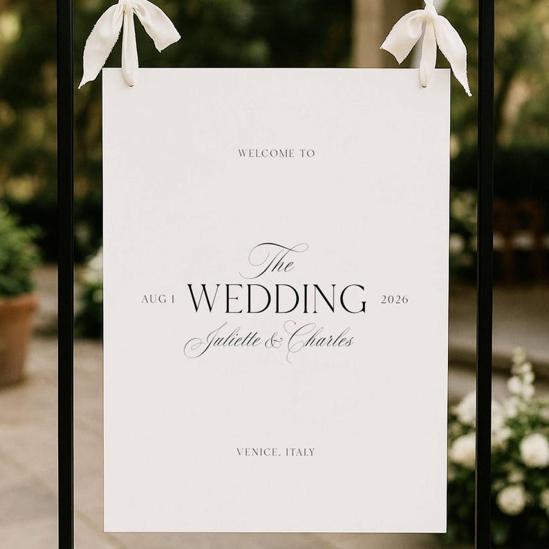 May include: A white wedding sign with black text, suspended from a black metal frame. The sign says "Welcome to The Wedding, Juliette & Charles, Aug 1, 2026, Venice, Italy." White ribbons are tied at the top.