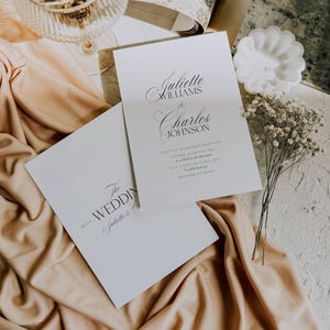 May include: Two white wedding invitations with elegant calligraphy. The top invitation reads "Juliette Williams & Charles Johnson." The bottom invitation reads "The Wedding." Soft peach fabric and delicate flowers accent the invitations.