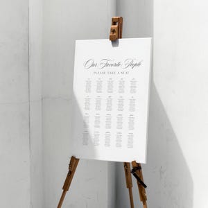 May include: A white seating chart with the text "Our Favorite People" and "Please Take a Seat" in elegant script. The chart is displayed on a wooden easel, perfect for a wedding or event.