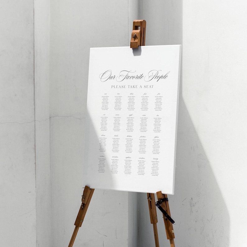 May include: A white seating chart with the text "Our Favorite People" and "Please Take a Seat" in elegant script. The chart is displayed on a wooden easel, perfect for a wedding or event.