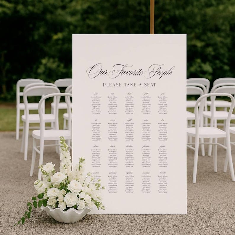 May include: A large cream-colored sign with the text "Our Favorite People PLEASE TAKE A SEAT" in elegant script. The sign lists names in columns. White chairs are arranged behind the sign. A white floral arrangement sits in front of the sign.