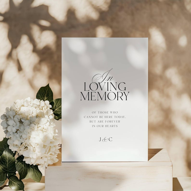 Op de afbeelding: Een wit bord met de woorden "In Loving Memory" in elegant zwart schrift. De tekst hieronder luidt: "Van degenen die er vandaag niet kunnen zijn. Maar voor altijd in onze harten." Links staat een boeket witte bloemen.
