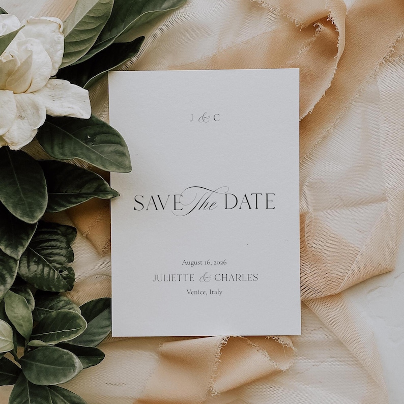 May include: A white Save the Date card with elegant script and the text "SAVE The DATE". The card includes the date August 16, 2026, and the names Juliette & Charles, with Venice, Italy, as the location. The card is set against a backdrop of soft fabric and greenery.