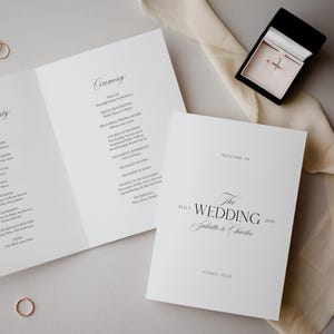 Puede incluir: Un conjunto de invitaciones de boda con un anillo en una caja negra, dos programas de boda y una alianza. El programa dice "Ceremonia" y "Bienvenida a la boda Juliette & Charles Venecia, Italia 1 de agosto de 2026."
