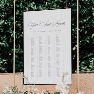 May include: A white seating chart with the text "Your Seat Awaits" in elegant script. The chart lists names alphabetically, with the names "Juliette & Charles" and the date "August 28, 2026" at the top. The chart is framed in black and suspended from a gold frame.