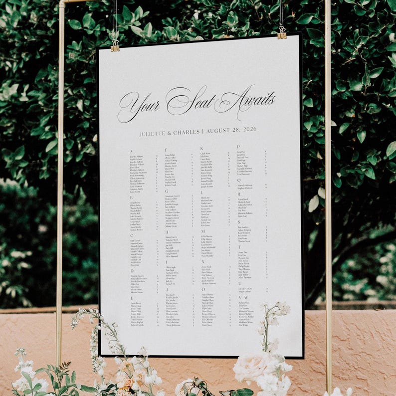 May include: A white seating chart with the text "Your Seat Awaits" in elegant script. The chart lists names alphabetically, with the names "Juliette & Charles" and the date "August 28, 2026" at the top. The chart is framed in black and suspended from a gold frame.