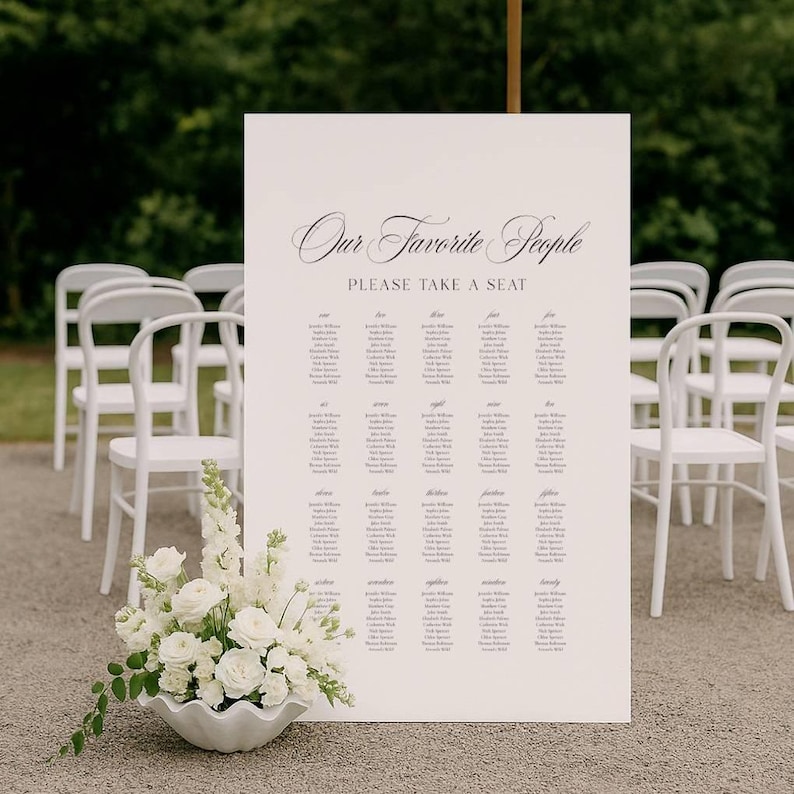 May include: An off-white sign with the text "Our Favorite People PLEASE TAKE A SEAT" in elegant script. Below, a list of names is organised in columns. White chairs are arranged in rows behind the sign, and a white floral arrangement sits in front.