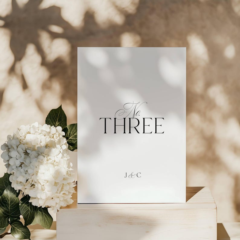 May include: A white card with the text "No. THREE" in elegant black script, with "J & C" below. The card is set on a light wooden surface, with white flowers and green leaves to the left, and shadows in the background.