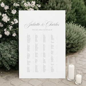 May include: A white rectangular wedding seating chart sign with black text reading Juliette & Charles and Please Find Your Seat, followed by an alphabetical list of names organized by letter columns, printed on a smooth, matte surface.