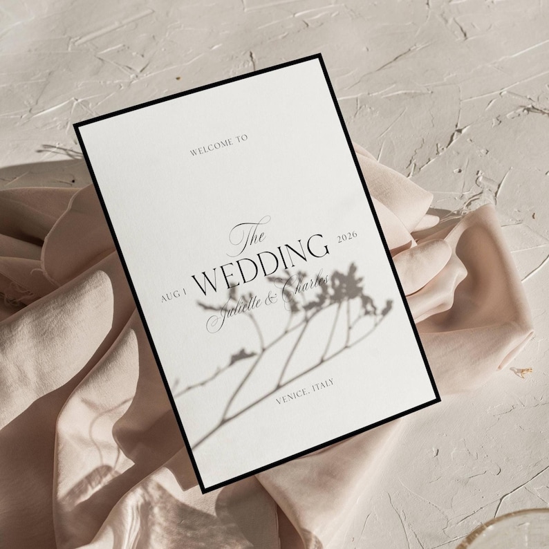 Pu&ograve; includere: Un invito di nozze con bordo nero, con le parole "Welcome to The Wedding" e i nomi "Juliette & Charles" con la data "1 agosto 2026" e la localit&agrave; "Venezia, Italia". L'invito &egrave; su sfondo bianco con un tessuto rosa tenue.