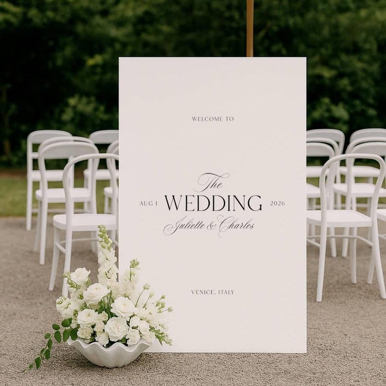 May include: A large white wedding sign with elegant script reading "Welcome to The Wedding, Juliette & Charles, Aug 1, 2026, Venice, Italy." White chairs are arranged behind the sign. A white floral arrangement sits at the base.