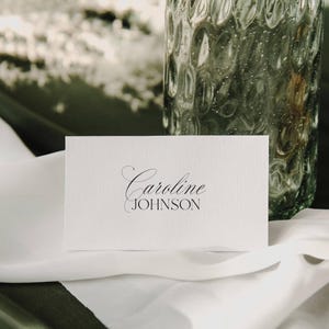 May include: A white place card with the name "Caroline Johnson" printed in elegant black script. The card is set on a white cloth, with a textured glass vase in the background.