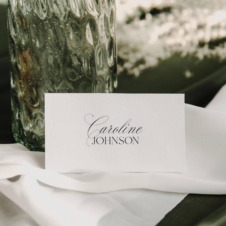 May include: A white place card with the name "Caroline Johnson" printed in elegant black script. The card is placed on a white cloth, with a textured glass vase in the background. The overall aesthetic is simple and sophisticated.