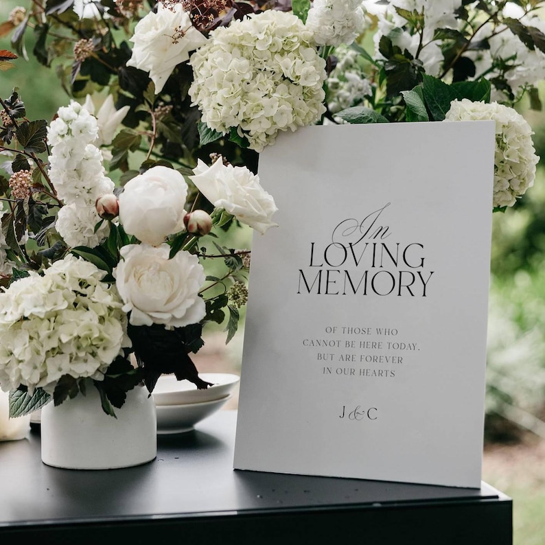Op de afbeelding: Een wit bord met de tekst "In Loving Memory" en een oprechte boodschap, tentoongesteld in de buurt van een bloemstuk van witte hortensia's en rozen. Het bord staat op een zwart oppervlak.