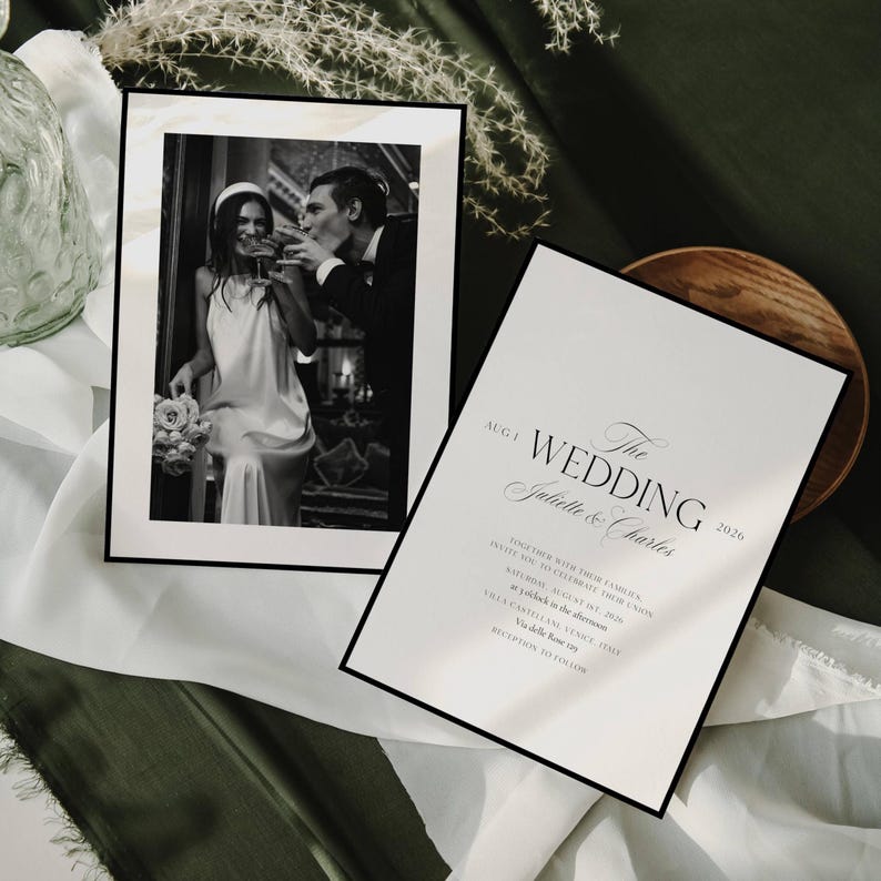 May include: A black and white photograph of a couple toasting, framed in black, next to a wedding invitation. The invitation is white with black text that reads "The Wedding Juliette & Charles 2026". The background is a mix of white and green fabric.