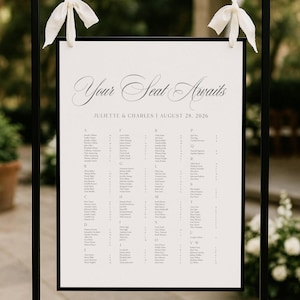 May include: A wedding seating chart with the words "Your Seat Awaits" in elegant script. The names of the guests are listed alphabetically. The chart is framed in black and adorned with white ribbons.