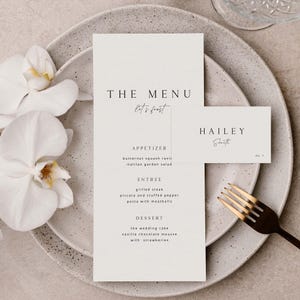 May include: A minimalist wedding menu and place card set. The menu features the text "THE MENU" and "Let's feast", with a list of courses. The place card reads "HAILEY Smith". A gold fork and white orchid complete the elegant table setting.