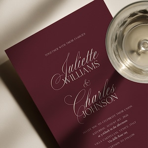 May include: Burgundy wedding invitation with elegant script lettering. The invitation reads "Juliette Williams & Charles Johnson" and includes event details for August 1st, 2026, in Venice, Italy. A glass of liquid is in the upper right corner.
