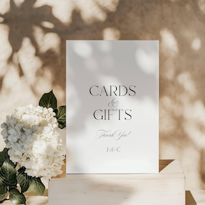 May include: A white sign with the words "CARDS & GIFTS" in elegant black script. Below, it reads "Thank You!" and initials "J & C". A bouquet of white flowers and green leaves sits to the left. The sign is on a light wooden surface.
