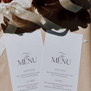 May include: Two white menu cards with elegant calligraphy reading "The MENU." The menu lists appetizers and entrees, including Caprese salad and herb-crusted chicken breast. A white ribbon and decorative elements are visible in the background.