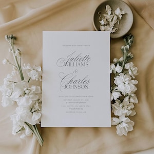 May include: A wedding invitation with the names Juliette Williams and Charles Johnson in elegant script. The invitation is surrounded by white flowers and rests on a beige fabric. The text invites guests to celebrate their union on Saturday, August 1st, 2026, in Venice, Italy.